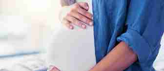 A close-up shot of a pregnant woman cradling her baby bump with both hands. She is wearing a blue shirt and white top, and is sitting down. The background is softly blurred, emphasizing the gentle and serene moment.