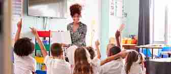 A teacher is seated at the front of a classroom, smiling and holding an open book. Several children wearing school uniforms are sitting on the floor in front of her, raising their hands enthusiastically. The classroom is colorful and well-lit.