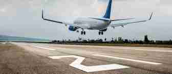 A commercial airplane with blue accents is landing on a runway under a partly cloudy sky, with trees and distant hills visible in the background.