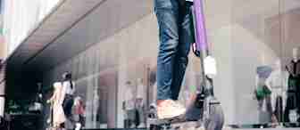 A person wearing a white shirt, blue jeans, and beige shoes rides a purple electric scooter on a city sidewalk. In the background, there are glass display windows showing mannequins, and two other pedestrians are walking. The person has a backpack on.