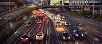 Evening scene of a busy urban highway with multiple lanes of vehicles under an illuminated overpass. A modern office building with bright lights stands prominently in the background against the twilight sky. Several vehicles have their headlights on.