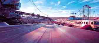 A conveyor belt runs through an open-pit mining site under a bright blue sky. Heavy machinery and cranes can be seen on either side, and rugged, rocky terrain stretches into the distance. A truck is parked to the right, emphasizing the scale of the operation.