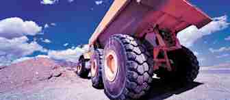 Low-angle view of a large orange dump truck with massive tires, positioned next to a pile of dirt under a vivid blue sky with scattered white clouds. The image captures the robust and heavy-duty nature of the construction vehicle.