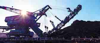 A large industrial excavator machine is working at a mining site. The sun shines brightly in the background, casting a glow on the machinery. Piles of excavated earth and rocks are visible beside the machine under a clear blue sky.