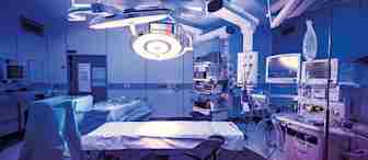 A modern operating room filled with medical equipment. There's a surgical table in the center, surrounded by monitors, overhead lights, and various machines. The lighting is dim with a bluish hue, giving an ambient atmosphere.