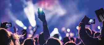 A lively concert scene where several attendees are holding up their smartphones to capture photos and videos. The stage is bathed in colorful lights, creating a vibrant atmosphere. The background is slightly blurred, with the focus on the raised phones.