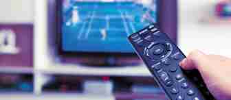 A hand holding a TV remote is pointed at a television screen showing a tennis match in a living room setting. Shelves with books and decorative items are visible in the background.