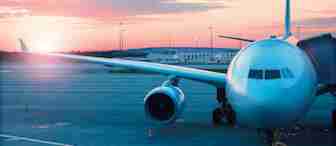 A large passenger airplane is parked at an airport gate during sunset. The sky features a mix of pink, orange, and blue hues while the sun is partially visible near the horizon. The airplane is connected to the gate by a jet bridge.