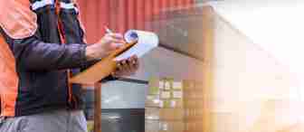 A person wearing a black and orange jacket holds a clipboard and writes notes. Behind them, a truck is being loaded with several stacks of cardboard boxes. The scene suggests logistics or delivery operations, with a focus on inventory or shipment documentation.