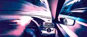 A blurred, high-speed view from inside a car, focusing on the driver's hand on the steering wheel, dashboard, and side mirror. The outside scenery and road ahead are motion-blurred, giving a sense of rapid movement and speed.
