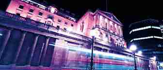 A historic building illuminated with pink and purple lights at night. Long exposure creates light trails from passing vehicles, enhancing the dynamic scene. Street lamps and modern buildings in the background contrast with the classical architecture.