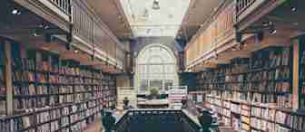 A cozy, two-story library with wooden bookshelves lining both sides and a central staircase leading to the upper level. The floor is wooden, and natural light pours in from a large, arched window in the background, illuminating the spacious interior.