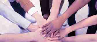 A group of diverse people standing in a circle place their hands on top of each other in the center, symbolizing unity and teamwork. The hands vary in skin tones and clothing styles, representing inclusivity and collaboration. The background is a light-colored floor.