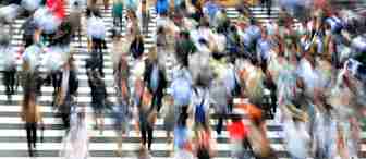 A blurred image of numerous people walking across a large, busy, zebra-striped crosswalk. The motion blur captures the hustle and bustle of the crowd, giving a sense of fast-paced urban life. The scene is vibrant with diverse clothing colors and patterns.