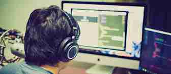 A person wearing headphones sits at a desk facing a large monitor that displays coding scripts. Another smaller monitor to the right also shows code. Various electronic devices and objects are on the desk. The scene suggests a focused work environment.