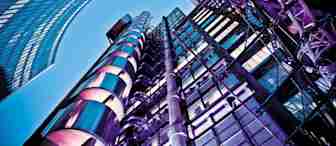 A striking upward view of modern skyscrapers with reflective glass and metal exteriors against a clear blue sky. The buildings are illuminated with purple and blue lights, showcasing a high-tech, futuristic architectural design.