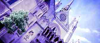 A low-angle view of an ornate Gothic-style building with pointed arches, intricate carvings, and a tall spire under a bright blue sky. A small green tree is visible on the left side of the building. The image has a purple hue.