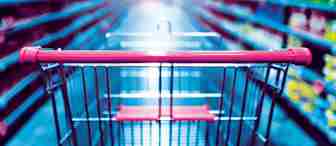 A shopping cart with a red handle is in focus in the foreground of a brightly lit supermarket aisle, surrounded by shelves stocked with a variety of products. The background is blurred, emphasizing the perspective from the shopper's point of view.