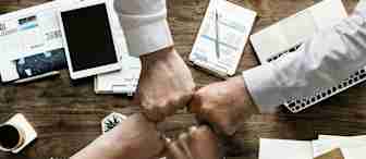 Four hands, each belonging to a different person, meet in a fist bump over a cluttered wooden desk. The desk holds various office items including papers, a tablet, a calculator, notebooks, a smartphone, and a cup of coffee.