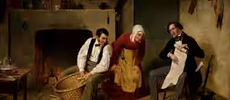 A cozy room with three people: a man shelling corn near a fireplace, a woman in a brown dress and white bonnet watching, and another man in a suit reading. The room is rustic and warmly lit, with various household items scattered around.