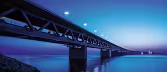 A long bridge illuminated by streetlights stretches across a calm body of water at twilight. The sky is a gradient of blue hues, and the bridge's reflection is visible on the water's surface. Rocks line the shore in the foreground.