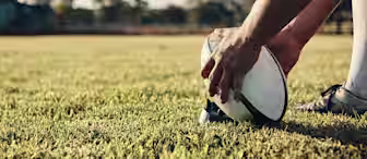 A rugby player, seen from the knees down, is setting a rugby ball on a kicking tee on a grassy field. The player is wearing white socks and cleats. The background is blurred with trees and a cloudy sky suggesting an open field or park location.