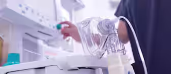 A medical professional in dark scrubs adjusts the settings on a ventilator machine. The foreground features the ventilator's control panel, tubing, and a clear oxygen mask resting atop the equipment.