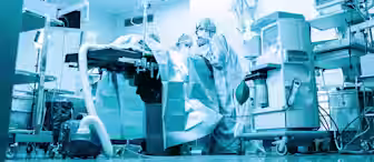 Medical team performing surgery in a modern operating room, surrounded by surgical equipment, monitors, and overhead lights; scene is tinted blue for a clinical effect.