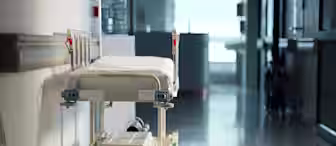 An empty hospital bed on wheels is positioned against the wall in a brightly lit corridor. The hallway appears quiet, with open space leading towards a distant door, suggesting an atmosphere of calmness in a medical setting.