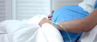 A pregnant woman wearing a blue hospital gown lies in a hospital bed with an IV drip attached to her hand. Her hands rest on her large belly, indicating she is in the later stages of pregnancy. The background shows white shutters.