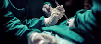 Surgeons in green scrubs and white gloves performing a surgical procedure. One surgeon holds a scalpel, and another assists. The scene is well-lit with focus on the patient's torso, while the background remains dark and out of focus.