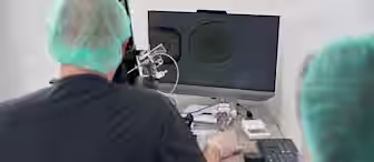 Two individuals in a laboratory setting are focused on a computer screen displaying a microscopic image. They are wearing surgical caps and gowns, indicating a sterile environment. The one on the left is manipulating a device, likely part of the microscope setup.