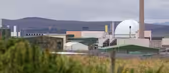 Industrial facility with a large dome-shaped structure and various buildings under a cloudy sky. In the foreground, there are grassy fields and fencing. Hills are visible in the background.