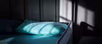 A neatly made bed with a light blue sheet and pillow is partially lit by sunlight streaming through a window, casting shadows. The room appears calm and tranquil.