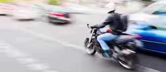 Blurred image of a motorcyclist wearing a helmet and backpack, riding swiftly through city traffic. Surrounding cars appear in motion, creating a dynamic and bustling urban scene.