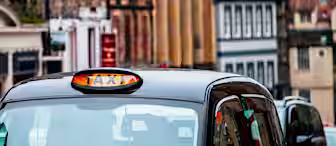 A black taxi with an illuminated "TAXI" sign on top is seen in a bustling urban street with blurred historic buildings in the background. Red flowers adorn a balcony, and a variety of architectural styles are visible.
