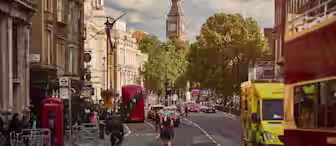 A bustling street in London with cyclists, cars, buses, and red phone booths. Big Ben is visible in the background under a partly cloudy sky. The street is lined with historic buildings and greenery. An ambulance is visible on the right side of the street.