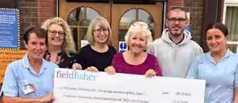 Six people, including three healthcare workers, smile while holding a large check in front of a hospital's Inpatient Unit. The check, from "Field Fisher," is for £23,731 and is made payable to the NHS for the Inpatient Unit.