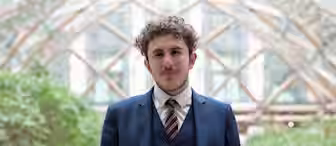 A person with curly hair and a beard is dressed in a blue suit with a white shirt and a striped tie. They are standing indoors with a blurred background featuring a geometric glass structure and greenery.