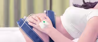 A pregnant woman lies on a bed while undergoing fetal monitoring. She is wearing a white outfit with blue straps around her belly connected to monitoring equipment, and has an intravenous line attached to her hand. Her other hand rests on her stomach.