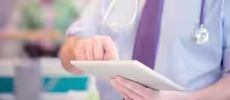 A healthcare professional wearing a shirt and tie, with a stethoscope around their neck, uses a tablet device. The background is blurred, showing another person in what appears to be a medical setting.