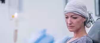 A woman with a headscarf, likely undergoing medical treatment, sits in a chair near an IV drip. She appears contemplative, highlighting the challenges faced by individuals receiving care. The setting is clinical, with blue chairs and a calm atmosphere in the background.