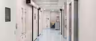 A well-lit, clean hospital corridor with light blue floors and white walls. It features sliding doors on either side and a small alcove with medical equipment and a chair at the end of the hallway. Fluorescent lights line the ceiling.