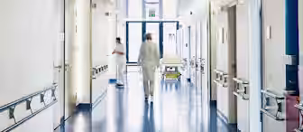 A brightly lit hospital corridor with white walls and shiny blue floors. A medical professional in white attire walks down the hallway toward a double door exit with windows. Another person, slightly blurred, stands near the wall on the left side.