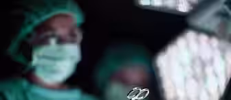 Yaser Jabbar, Orthopaedic Surgeon at Great Ormond Street Hospital - picture of Surgeons in sterile green gowns and masks are performing a surgery. One surgeon is passing a pair of surgical scissors to another. The scene is illuminated by surgical lights, with a focus on the scissors and the gloved hands.