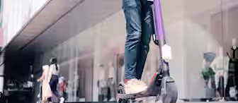 A person wearing a white shirt, blue jeans, and beige shoes rides a purple electric scooter on a city sidewalk. In the background, there are glass display windows showing mannequins, and two other pedestrians are walking. The person has a backpack on.