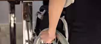A person seated in a wheelchair gripping the wheel rim. They are partially visible, wearing a white shirt and black pants, positioned near a shiny, reflective table. Sunlight illuminates the scene, highlighting the hand's detail and wheelchair's structure.