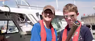 Two people wearing red life jackets and sunglasses smile on a boat. One holds a metal object while the other places a hand on their shoulder. The background shows a sunny marina with boats and a pier.