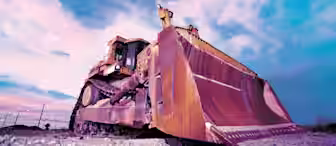 A low-angle shot of a large bulldozer with a wide blade on a construction site under a dramatic blue and pink sky. The earth around the bulldozer appears partially leveled, indicating ongoing construction work.