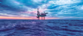 An offshore oil rig stands tall in the middle of a vast ocean, under a dramatic sky filled with clouds at sunset. The platform is surrounded by deep blue water, with the horizon displaying shades of orange and purple as the day transitions to night.